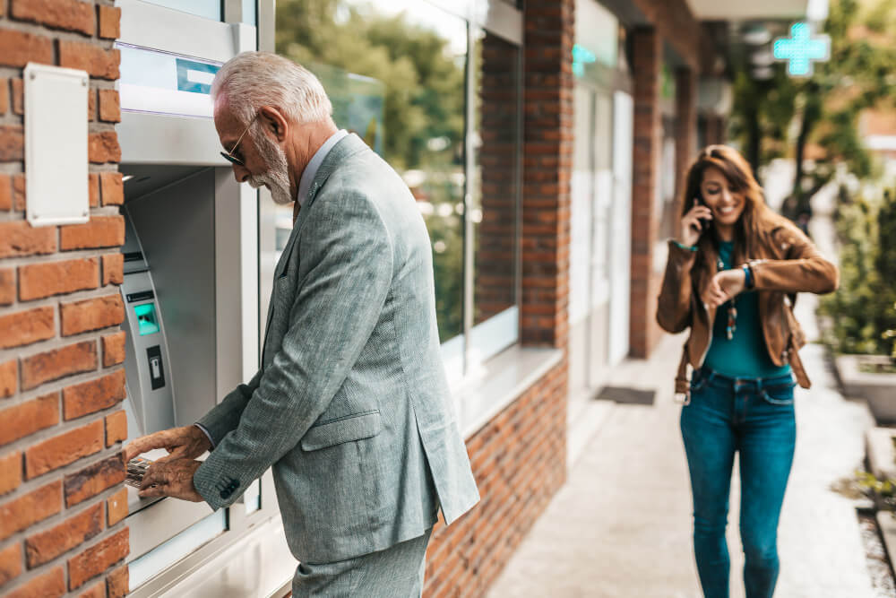 A man withdrawing money from an atm with a woman walking by
